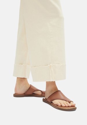 Braune Leder-Slidesandalen mit minimalistischen Design, ausgestattet mit einem breiten Riemen und einer flachen Sohle, kombiniert mit cremefarbigen, cropped Hosen.