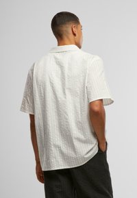 Camicetta a maniche corte in tessuto beige chiaro con una trama ondulata e testurizzata, caratterizzata da un colletto classico e una vestibilità informale.