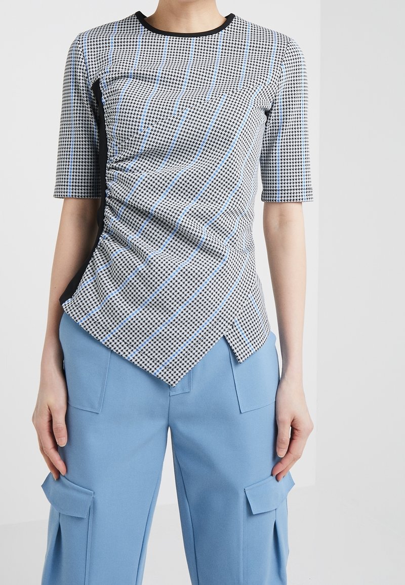 Haut à carreaux noirs et blancs avec des rayures bleues, manches courtes et ourlet asymétrique. Le tissu est lisse avec une texture subtile.