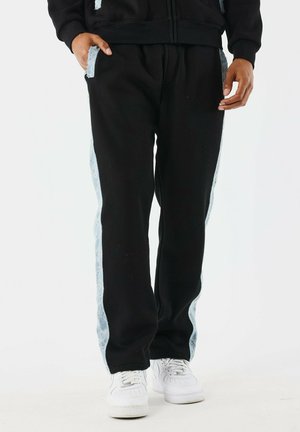 Pantaloni neri con pannelli laterali azzurri, vita elasticizzata e tasche, realizzati in un tessuto morbido. Indossati con sneakers bianche.