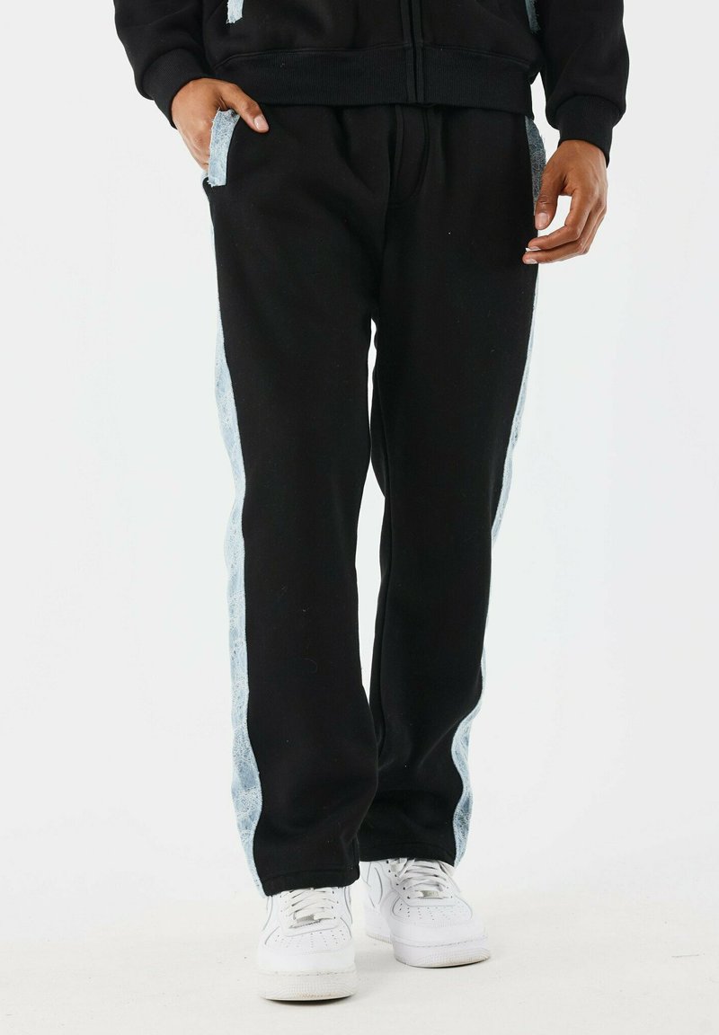 Pantalones de chándal negros con paneles laterales azul claro, cintura elástica y bolsillos, fabricados en una tela suave. Se combinan con zapatillas blancas.