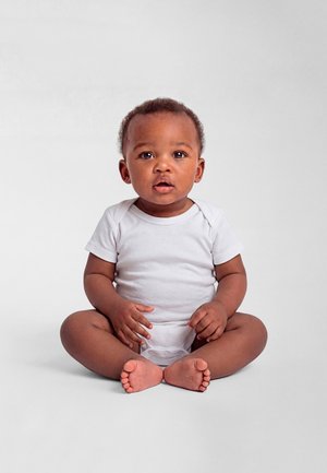 Rövid, göndör hajú baba ül keresztbe tett lábakkal egy egyszerű fehér felületen, fehér rövid ujjú bodyt visel, előre néz, szája kissé nyitva.