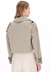 Kurz geschnittener beiger Trenchcoat mit weitem Kragen, Schulterklappen und Knopfakzenten. Glatter Stoff mit tailliertem Schnitt und Manschetten-Beschlägen.