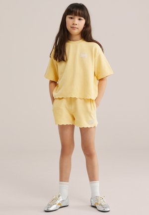 Jeune fille debout les mains dans les poches, portant un t-shirt et un short jaunes assortis à bords festonnés, des chaussettes blanches et des baskets argentées.