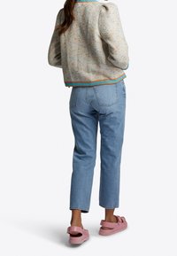 Veste légèrement texturée, multicolore, avec des manches bouffantes et des accents bleus, associée à un jean bleu clair et des sandales roses. Vue arrière décontractée.