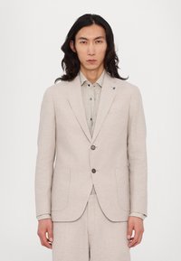 Veste de costume en lin beige avec un design structuré, deux boutons et deux poches avant. Portée sur une chemise à col gris clair.