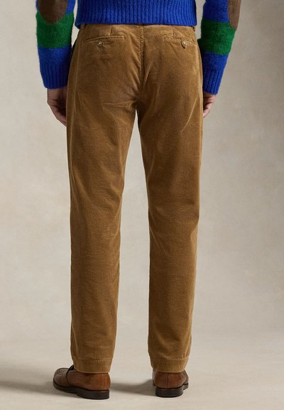 Man die bruine corduroy broek, bruine leren schoenen en een blauwe trui met groene en bruine elleboogstukken draagt, staat met zijn rug naar de camera.