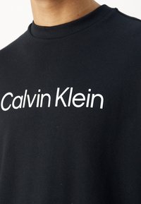 Sweat-shirt noir avec un col côtelé et un logo blanc audacieux "Calvin Klein" sur la poitrine. Tissu en coton doux avec une texture lisse.