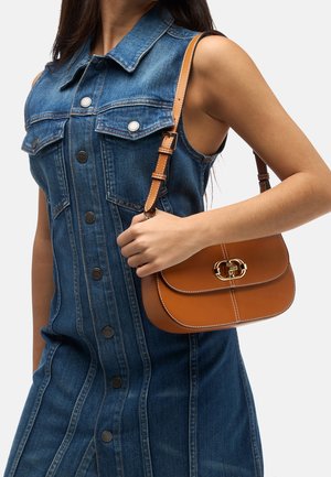 Kobieta w niebieskiej, bezrękawowej sukience dżinsowej, trzymająca na ramieniu brązową skórzaną torebkę z złotym zapięciem.