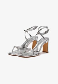 Silver metallic high-heeled sandals with square toe, thin straps, and a block heel. Features a smooth texture and adjustable ankle strap.