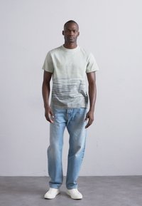 Utvasket grønn t-skjorte med horisontale grå striper, kombinert med lyseblå jeans og hvite sneakers, stående mot en nøytral bakgrunn.