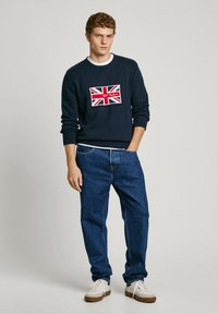 Navy sweater with white crew neck, featuring Union Jack design. Blue jeans and white sneakers complete the outfit. Simple, casual style.