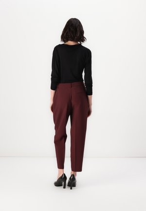 Vrouw met kort donker haar staat voor een witte muur, draagt een zwarte top met lange mouwen, bordeauxrode broek en zwarte hoge hakken.