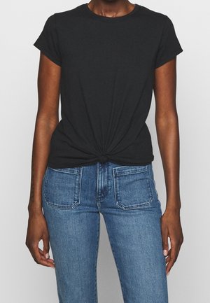 Persoon draagt een zwart T-shirt met korte mouwen, vooraan geknoopt, gecombineerd met high-waisted blauwe spijkerbroek met zichtbare voorzakken.