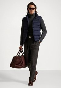 Granatowa pikowana kamizelka na ciemnoszarym swetrze, w połączeniu z szarymi spodniami. Nosi brązową skórzaną torbę podróżną, okulary przeciwsłoneczne i brązowe buty.