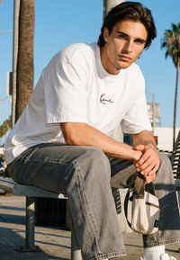 Mlad muškarac u bijeloj majici i sivim trapericama sjedi na metalnoj klupi držeći malu bež i crnu torbu na otvorenom kraj palmi.