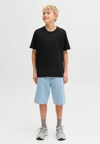 Crna pamučna majica kratkih rukava s natpisom "ORIGINALS" ispisanim u suptilnom fontu, uparena s svijetloplavim trapericama, sivim čarapama i sivim sportskim cipelama.