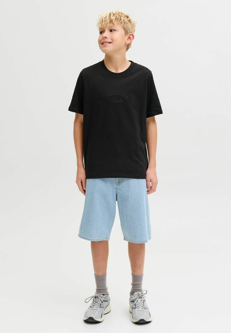 Crna pamučna majica kratkih rukava s natpisom "ORIGINALS" ispisanim u suptilnom fontu, uparena s svijetloplavim trapericama, sivim čarapama i sivim sportskim cipelama.