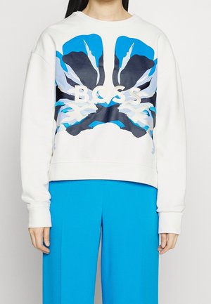 Sweat-shirt blanc avec de grandes ailes de papillon abstraites bleues et noires et le mot "BOSS" sur la poitrine, porté avec un pantalon bleu vif.