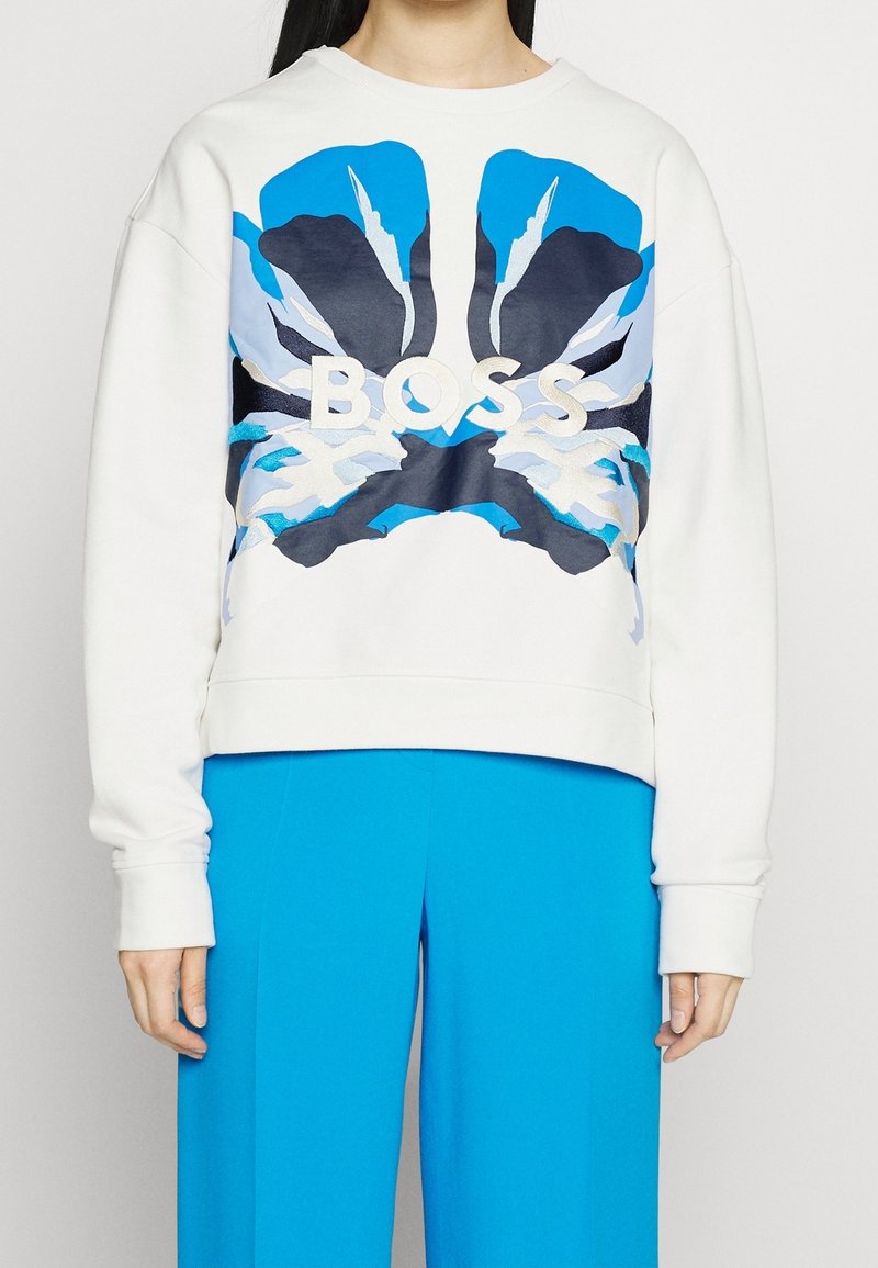 Sweat-shirt blanc avec de grandes ailes de papillon abstraites bleues et noires et le mot "BOSS" sur la poitrine, porté avec un pantalon bleu vif.