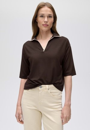 Vrouw draagt een donkerbruin poloshirt met korte mouwen en een crème randje bij de kraag, en lichtbeige broek, staand voor een effen lichte achtergrond.