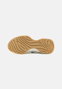 Semelle en caoutchouc marron clair avec motif ondulé et texturé, et logo circulaire près du talon sur fond blanc.