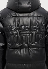 Crna puffer jakna s glatkom kožnom završnom obradom, s ugraviranim tekstom "VERSACE JEAN COUTURE" na leđima i kapuljačom.