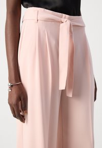 Pantaloni rosa pallido a vita alta con pieghe, una cintura in tessuto e una texture liscia. Presentano una tasca laterale.