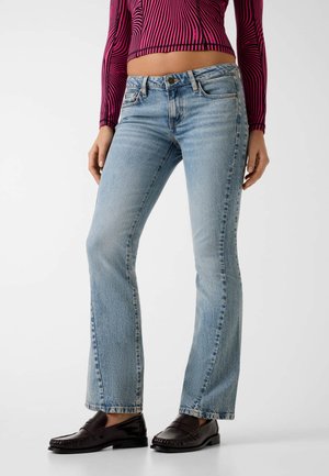 Vrouw draagt lichtblauwe flared jeans, zwarte loafers en een cropped longsleeve top met rode en zwarte golvende strepen.