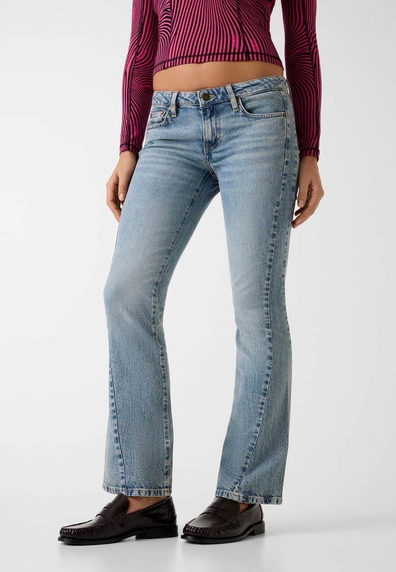 Femme portant un jean flare bleu clair, des mocassins noirs et un haut court à manches longues avec des rayures ondulées rouges et noires.