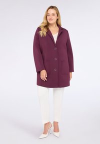 Cappotto bordeaux con colletto a punta, due tasche frontali, grandi bottoni e vestibilità rilassata, abbinato a pantaloni bianchi e scarpe con tacco.