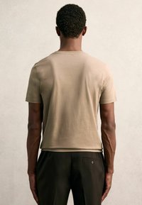 Bēšas krāsas īsām piedurknēm T-krekls no mīksta auduma, ar klasisku apaļu kakla izgriezumu un brīvu piegriezumu. Valkāts kopā ar tumšām biksēm.