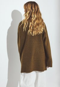 Olive green, oversized knit sweater with long sleeves and ribbed cuffs. Soft texture, drop shoulders, and a straight hem. White trousers below.