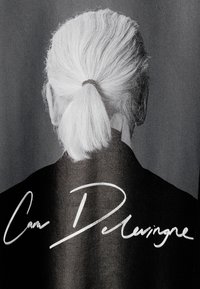 Černobílý obrázek zobrazující záda osoby s dlouhými, bílými vlasy stylizovanými do copu, oblečené v černém tričku s podpisem.