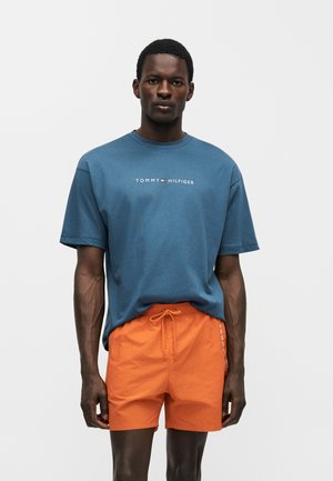 Man wearing blue Tommy Hilfiger t-shirt and orange drawstring shorts, standing against plain light background.