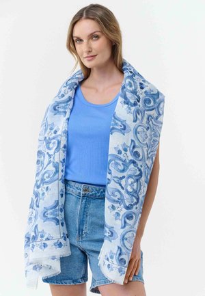 Woman wearing a blue tank top and denim shorts, draped with a white scarf featuring blue floral and swirl patterns.