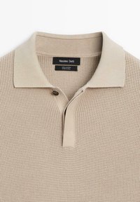 Polo en coton beige avec un tissu texturé, un col plat, une patte à deux boutons et une petite étiquette de marque au niveau du col.