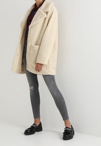 Persona che indossa un cappotto teddy crema, una camicia a quadri rossi, jeans skinny grigi strappati e mocassini neri con catene argentate, in piedi contro un muro bianco.