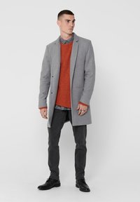 Grau übermantel aus dickem Stoff, getragen über einem orangefarbenen Pullover und einem grauen Hemd. Schwarze Slim-Fit-Hose und schwarze Schuhe runden das Outfit ab.