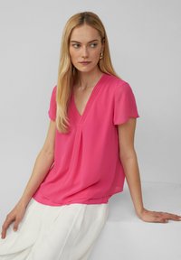 Kurzärmeliges V-Ausschnitt-Top aus lebhaft pinkfarbenem Stoff mit Falten an der Vorderseite. Glatte Textur mit einer lockeren Passform, kombiniert mit weißen Hosen.