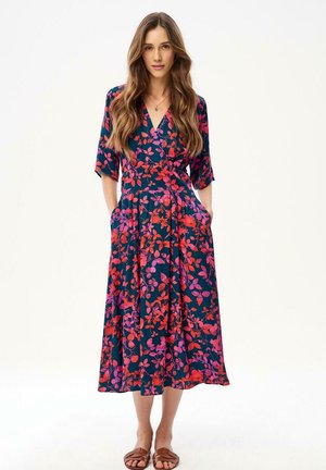 Kobieta w granatowej sukience midi z czerwono-różowymi kwiatowymi wzorami, z rękawami do łokcia i brązowych płaskich sandałach, stojąca z rękami w kieszeniach.