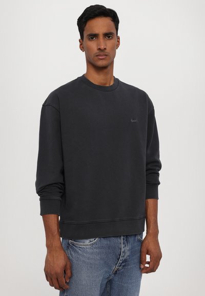 Sweatshirt charbon avec col rond, poignets et ourlet côtelés. Présente un petit logo sur la poitrine et une coupe décontractée. Porté avec un jean bleu.