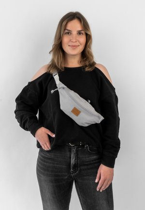 Grau gefärbte Stoffgürteltasche mit einem vorderen Reißverschluss, einem braunen Lederbesatz, verstellbarem Gurt und einer glatten Textur, über einem schwarzen Sweatshirt getragen.