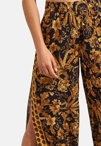 Pantalons en tissu noir avec un motif floral et de feuilles dorées, dotés d'une taille froncée et de fentes latérales pour faciliter les mouvements.