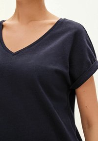 Námořnická modrá bavlněná tričko s výstřihem do V, krátkými ohrnutými rukávy a uvolněným střihem. Textura látky se zdá být hladká a lehká.