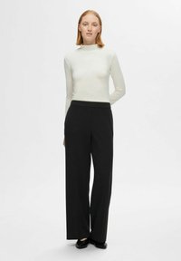 Weißes Langarmoberteil mit hohem Kragen, kombiniert mit schwarzen, weit geschnittenen Hosen. Glatter Stoff, minimalistisches Design und eine taillierte Silhouette.
