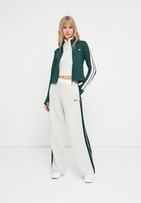 Zelena jakna na zip s bijelim prugama, uparena s svijetlosivim hlačama širokih nogavica koje imaju zelene bočne pruge i logo. Bijele cipele upotpunjuju look.