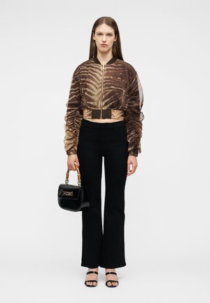 Femme debout portant une veste courte marron à motifs, un pantalon noir évasé, des sandales noires, et tenant un sac à main noir avec des poignées en bois.