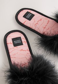 Sorte tøfler i imiteret pels med en pink inderside med et rødt krusedmønster og en rektangulær label med teksten "ROCKETT st GEORGE."