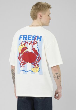 Jeune homme portant un t-shirt blanc avec un grand crabe rouge et un citron imprimés ainsi que le texte "FRESH crab" dans le dos, vu de côté.
